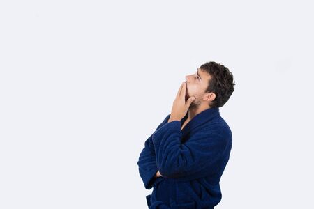 Sleepy young businessman yawning wearing blue bathrobe looking up,  feel tired needs to sleep isolated on white background.の写真素材