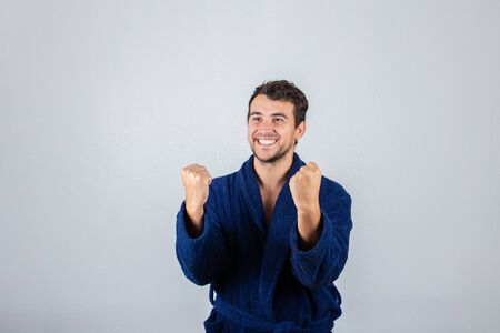 Portrait of cheerful young man, positive smiling  raising fists in victory or win gesture wearing blue bathrobe, isolated over white background.の写真素材