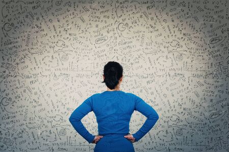 Rear view of a puzzled woman in front of a wall, hands on hips, try to solve hard mathematics...