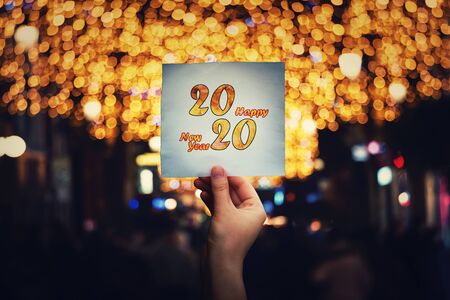 Hand holds a paper sheet with 2020 happy new year text message in the middle of a crowded night street with different Christmas lights and decorations ambiance. Winter holidays celebration concept.の写真素材
