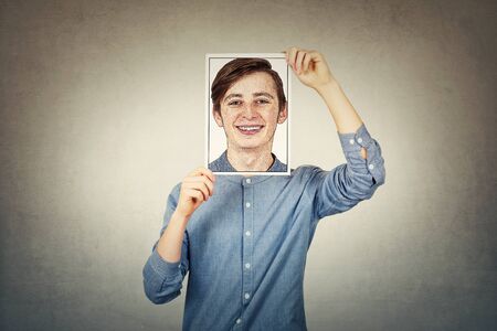 Teenage boy covering face using a paper photo print with self portrait sketch, like a mask for hiding the true emotion. Private life, dissembler false personality and fake identity concept.の写真素材