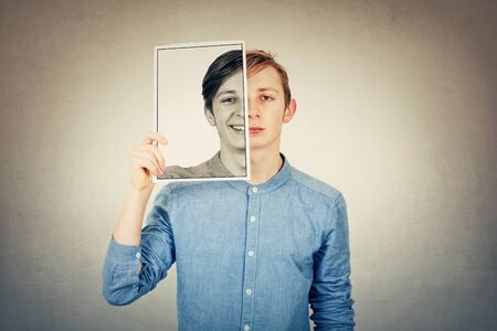 False portrait of a sad boy teenager hiding half face using a photo print paper sheet with a fake happy emotion. Mask for hiding the real expression, create new fake identity, grey wall background.の写真素材