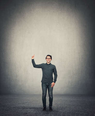 Full length contented businessman pointing forefinger up, standing in a room with gray wall background, empty copy space above head. Joyful business person wearing eyeglasses, shows index upwards.の写真素材