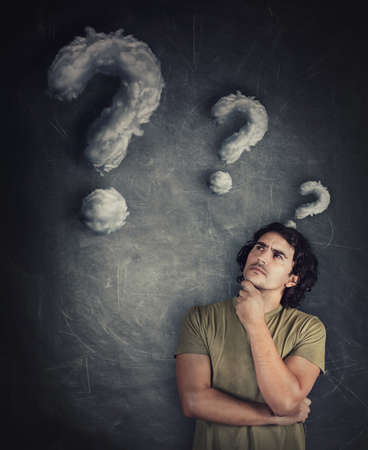Confused young man, curly hair, keeps hand under chin, thoughtful gesture, looking suspicious aside as cloud shaped interrogation marks leaving his head. Puzzled guy, has many questions, quiz concept.の写真素材