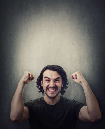 Pleased and positive man, long curly hair style, celebrates success hysterical and passionate. Guy keeps fists tight, raise hands up clenching teeth, isolated on gray wall with copy space above headの写真素材