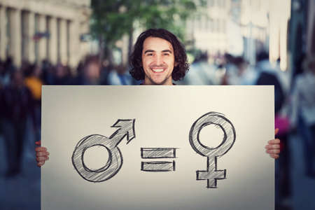 Gender equality concept as young man holding a big banner with male and female symbol, as protest demonstration message on a crowded city street. Sex rights and discrimination as a major social issueの写真素材