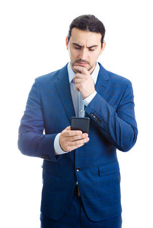 Thoughtful businessman keeps hand under chin, looking at his phone isolated on white background. Reflective business person planning and thinking for genius ideas.の写真素材