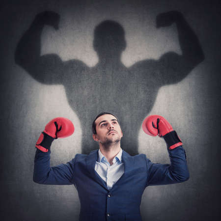 Confident businessman in suit with red boxing gloves celebrating success, casting a shadow of muscular and powerful leader on the wall. Flexing arm muscles, victory and business leadership conceptの写真素材