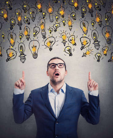 Amazed businessman pointing up index fingers, looking shocked as multiple light bulb float over his head. Impressed business person choosing from different creative ideasの写真素材