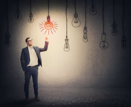 Cheerful businessman points up index finger, showing an enlightened light bulb among multiple others switched off. Business metaphor, idea choice conceptの写真素材