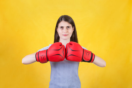 Young woman with red boxing gloves stands ready in a fighting stance, punching her fists. Portrait of determined girl prepared for battle isolated on yellow background. Self defense conceptの写真素材