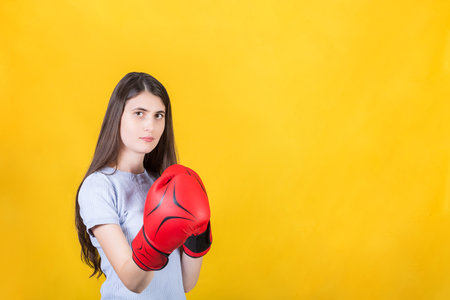 Confident young woman with red boxing gloves stands in fighting stance. Portrait of determined girl ready for battle isolated on yellow background with copy space. Self defense and leadership conceptの写真素材