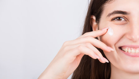 Young woman face closeup, using softening cream. Beautiful girl spreading moisturizer on her cheek, isolated on white background with copy space. Beauty skincare, cosmetics, spa procedures conceptの写真素材