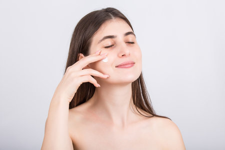 Young woman using softening face cream. Beautiful girl spreading moisturizer on her cheek, isolated on white background. Beauty treatment, skincare concept. Applying cosmetics, spa proceduresの写真素材