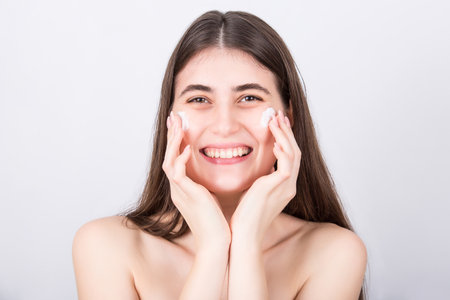 Happy young woman spreads moisturizing face cream on her cheeks. Beautiful girl apply softening moisturizer, isolated on white background. Skincare, cosmetics, beauty spa procedures conceptの写真素材