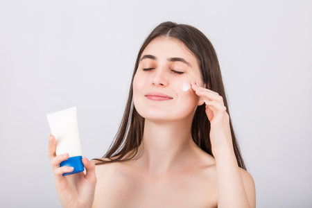 Beautiful woman applying softening face cream as holds a tube with moisturizing cosmetics in her hand, isolated on white background. Skincare concept and treatment spa procedures marketing and advertの写真素材