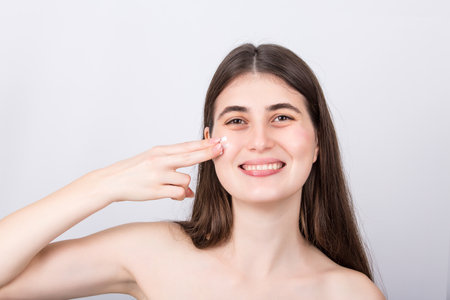 Excited young woman spreads moisturizing cream on her cheek, looking cheerful to camera, isolated on white background. Skincare, face cosmetics, beauty spa procedures conceptの写真素材
