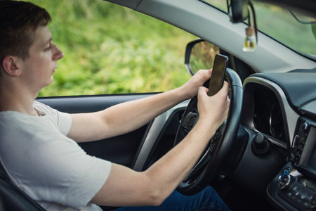 Young driver guy distracted by his phone while in front of the steering wheel, using his smartphone...