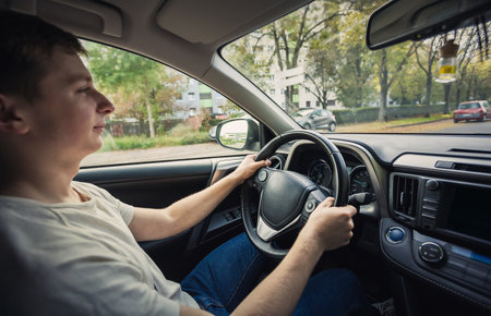 Contented young man driving car confident. Teenager driver enjoying the ride on the city streets, keeps hands on the steering wheel looking ahead at trafficの写真素材