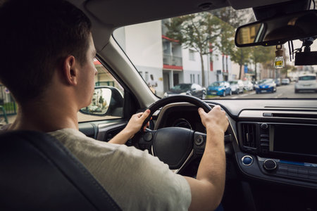 Confident young man driving car on the city streets. Teen driver keeps hands on the steering wheel looking ahead at the urban trafficの写真素材