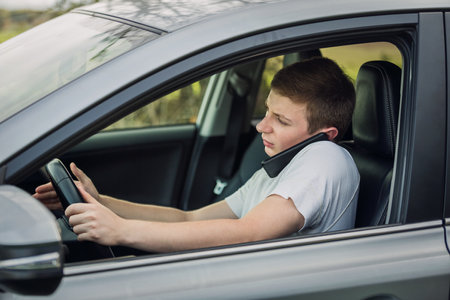 Young guy unsafe driving while talking on his phone in front of the steering wheel. Driver using his smartphone. Risk and danger situations on the road, violating traffic rulesの写真素材