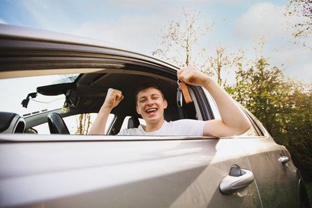 Happy Novice driver keeps fist up tight looking out of the driver window of his new car holding key from a new car. Successful young man bought a new car, celebrating triumph. Fun road trip.の写真素材