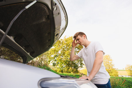Stressed novice driver looking at the car engine, hand to forehead gesture. Vehicle breakdown concept, automobile road problems. Open hood damaged motor.の写真素材