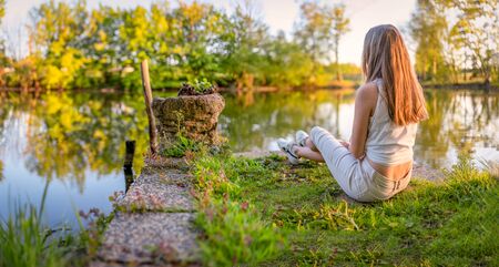 Young, caucasian girl sits relaxed by a pond in the evening sun and relaxesの写真素材