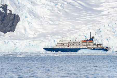 A blue, small expedition ship lies in front of an Antarctic iceberg landscape in Cierva Cove - a deep inlet on the west side of the Antarctic Peninsula, surrounded by rugged mountaの写真素材
