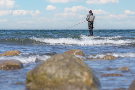 An angler in waterproof trousers is fishing in the Baltic Sea. He is standing on a stone in the water - Sunset on the Baltic Sea with a lone fisherman in the waterの写真素材