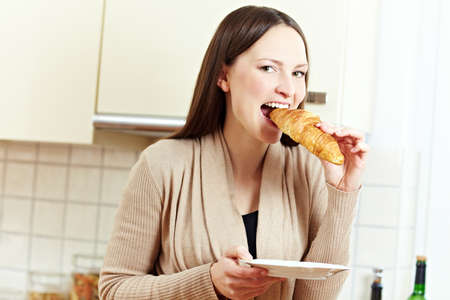 Happy woman biting in a croissant in a kitchenの写真素材