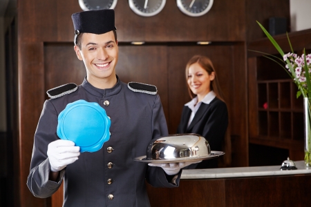 Friendly bellboy holding empty sign in hotel with receptionist behind counterの写真素材