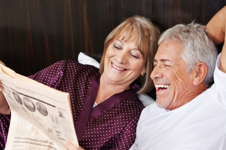Happy senior couple reading newspaper together in bedの写真素材