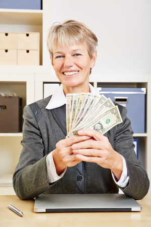 Elderly happy business woman with fan of dollar bills in the officeの写真素材