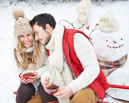 Happy couple with dog drinking tea in winter sitting on a sled