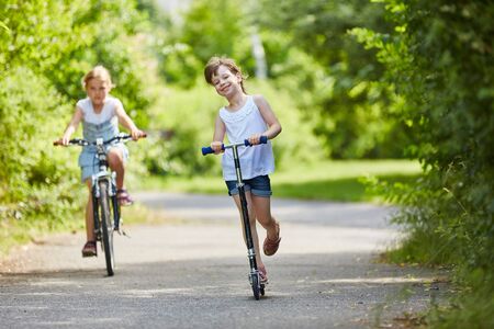 Girls drive scooter and ride bike in the park during summer vacationの写真素材