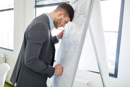 Frustrated businessman with head on whiteboard in officeの写真素材