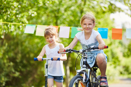 Two children riding bycicle and scooter in summer in the parkの写真素材