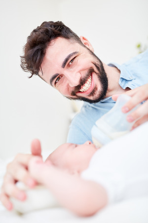 Happy husband as father on parental leave gives his baby milk from the bottleの写真素材