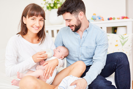 Happy parents give the bottle with milk to their baby at homeの写真素材