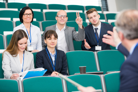 Confident businessman raising hand while colleague giving presentation in lecture hall