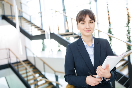 Portrait of confident businesswoman holding documents while standing against staircases in office