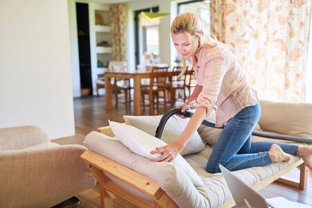 Cleaning lady with vacuum cleaner vacuuming a sofa in the living room