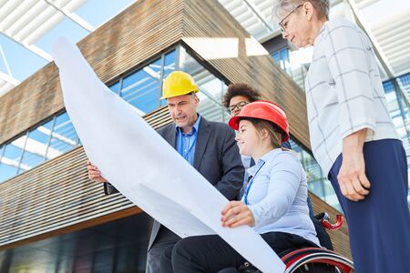 An engineering team with a blueprint and an architect in a wheelchair are planning an urban development projectの写真素材
