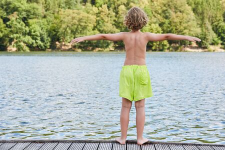 Little boy in shorts stands on a jetty at the bathing lake with outstretched armsの写真素材