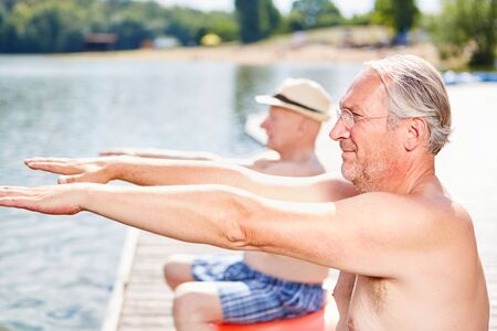 Two senior men stretch their arms during back exercises on the lake in summerの写真素材