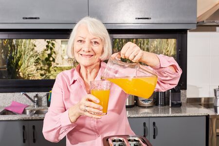 Smiling senior citizen pours orange juice from a carafe into a glass in the kitchen