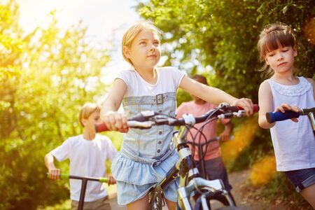 Children ride together on bicycles and scooters in summerの写真素材