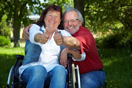 Laughing elderly woman in a wheelchair and her husband hold their thumbs up