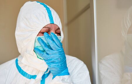 Tired doctor in protective clothing yawning holds hand in front of face in clinic during coronavirus pandemicの写真素材